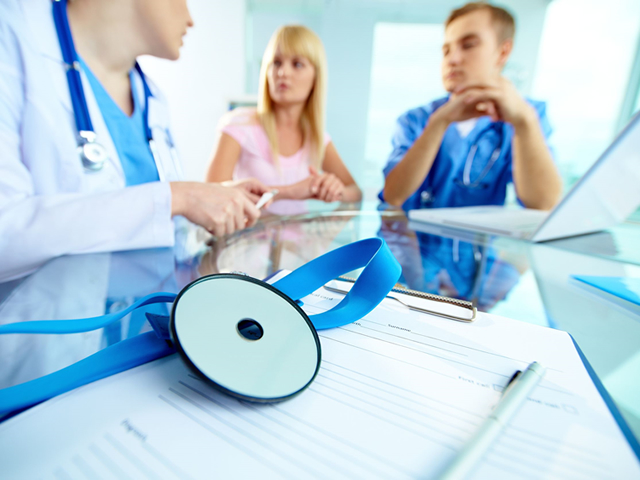 A stethoscope sits on top of a clipboard on a table as a doctor talks to a couple in the background